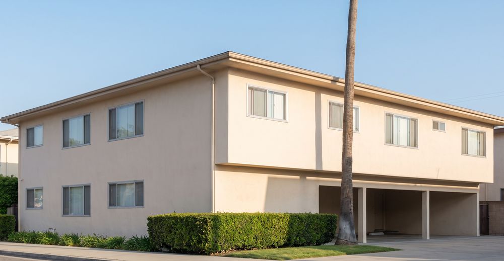 A two-story stucco apartment building with carport parking under the upper floor and a palm tree out front